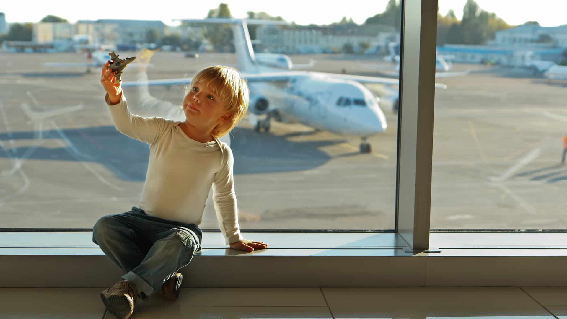 little boy sitting in airport playing with a toy plane with a plane behind him on the parking terminal
